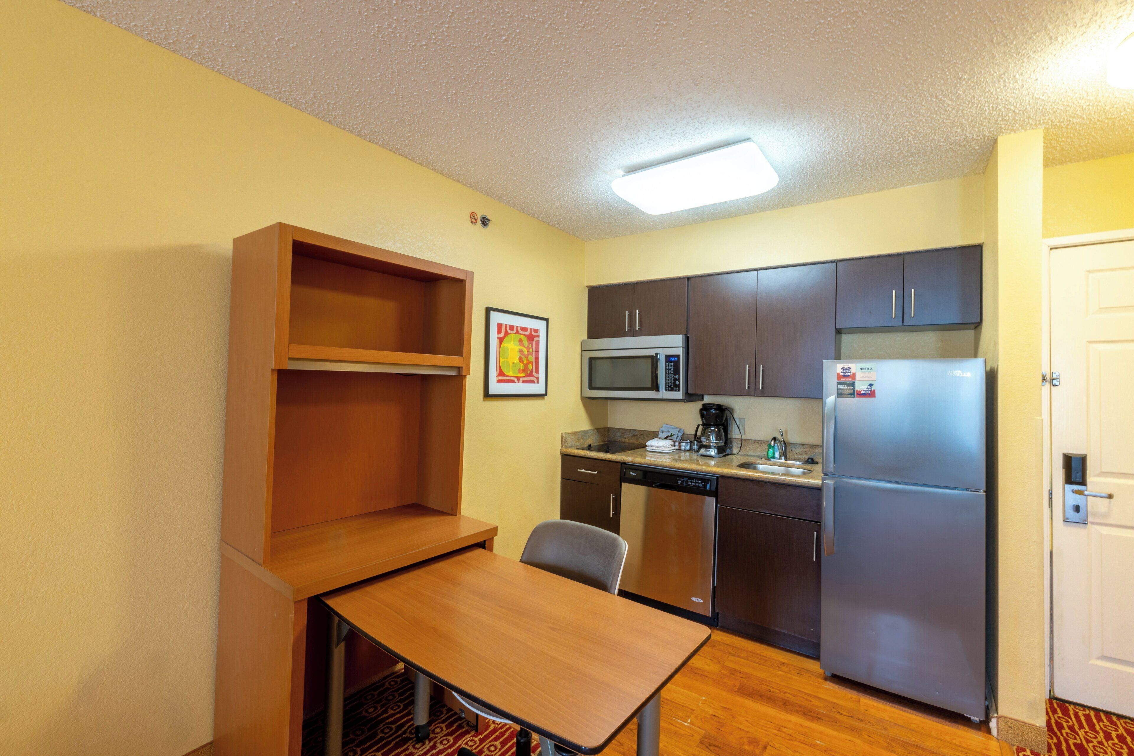 A compact kitchen with a microwave, refrigerator, and dishwasher. There's a small table and a chair, and a shelf unit is visible.