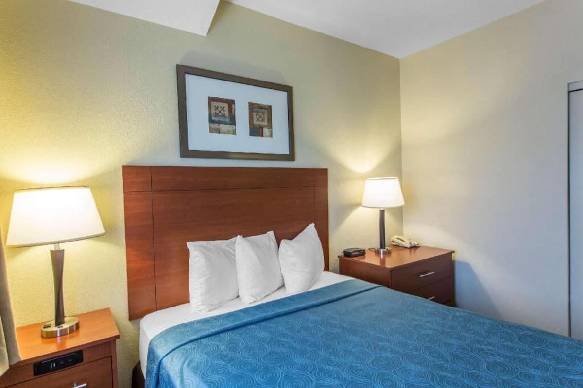 Habitación de hotel con cama, dos mesitas de noche, un reloj de pared y un cuadro enmarcado sobre la cama.