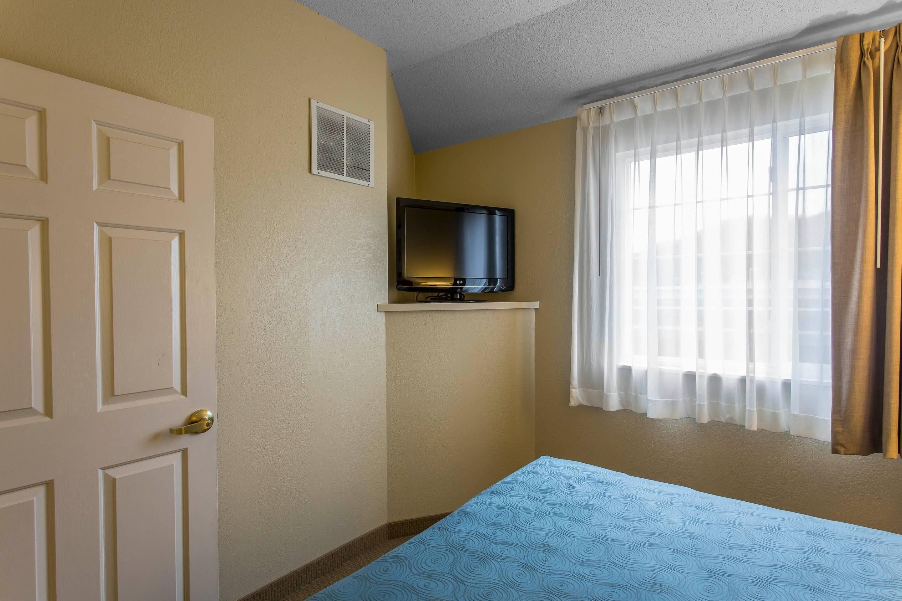 Una habitación con una cama, un televisor sobre un mueble bajo, una puerta y una ventana con cortinas.