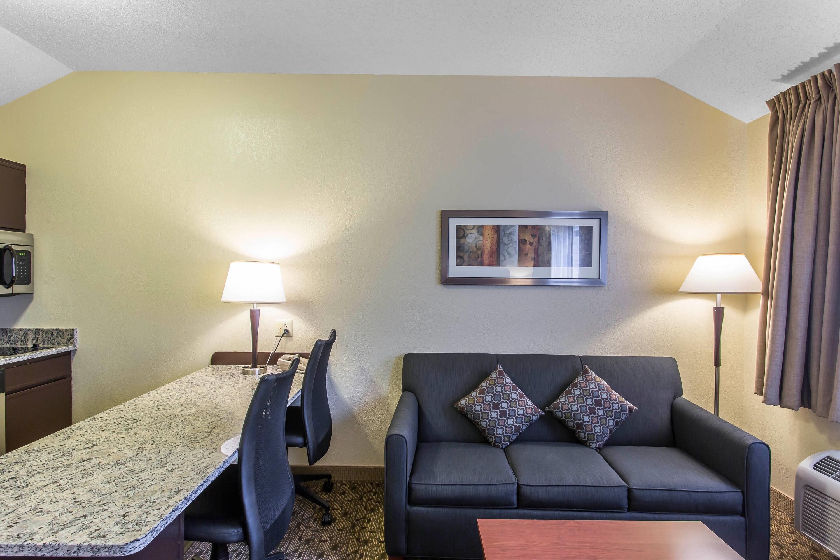 Habitación de hotel con una mesa de comedor, dos sillas, un sofá con cojines estampados, un cuadro en la pared y una lámpara de pie.