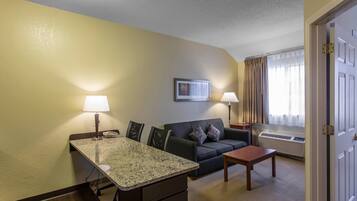 Habitación de hotel con mesa de comedor, sofá, pequeña mesa, lámpara, un cuadro en la pared y ventana con cortinas.