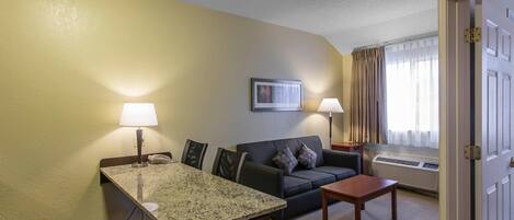 Habitación de hotel con mesa de comedor, sofá, pequeña mesa, lámpara, un cuadro en la pared y ventana con cortinas.