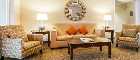 A hotel lobby with a patterned carpet, a sofa with orange pillows, two patterned armchairs, a coffee table, and a decorative mirror on the wall.