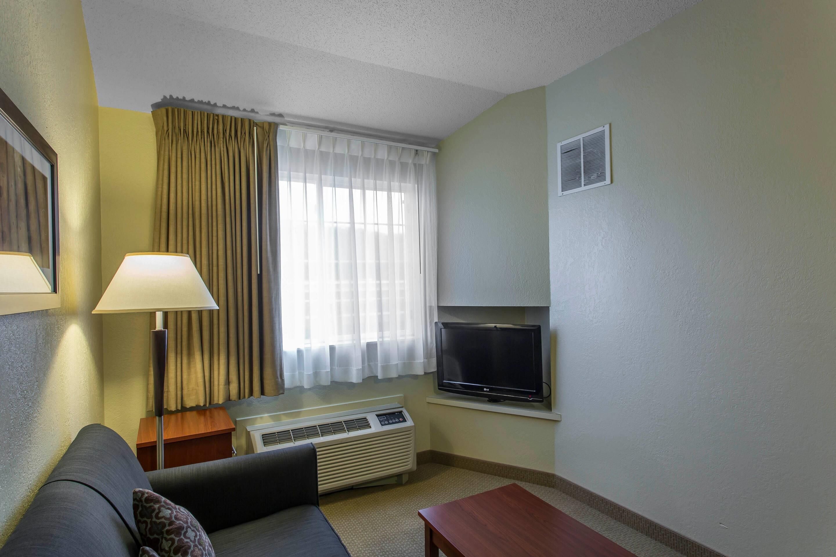 Una habitación de hotel con sofá, mesa, televisión, lámpara y ventana con cortinas.