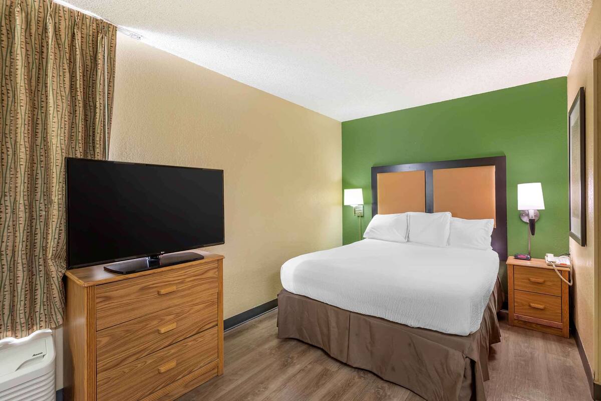 Ein Hotelzimmer mit einem Bett, einem Fernseher auf einer Holzkommode, einem Nachttisch mit Lampe und einer grünen Akzentwand.