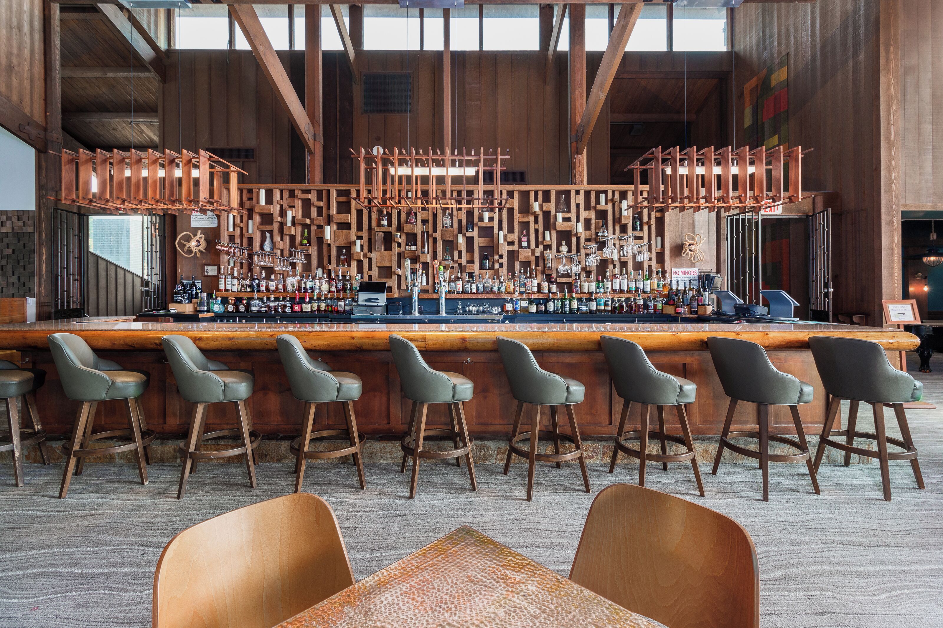 Una zona de bar con taburetes de madera, una barra con varias botellas y un techo alto con vigas a la vista.