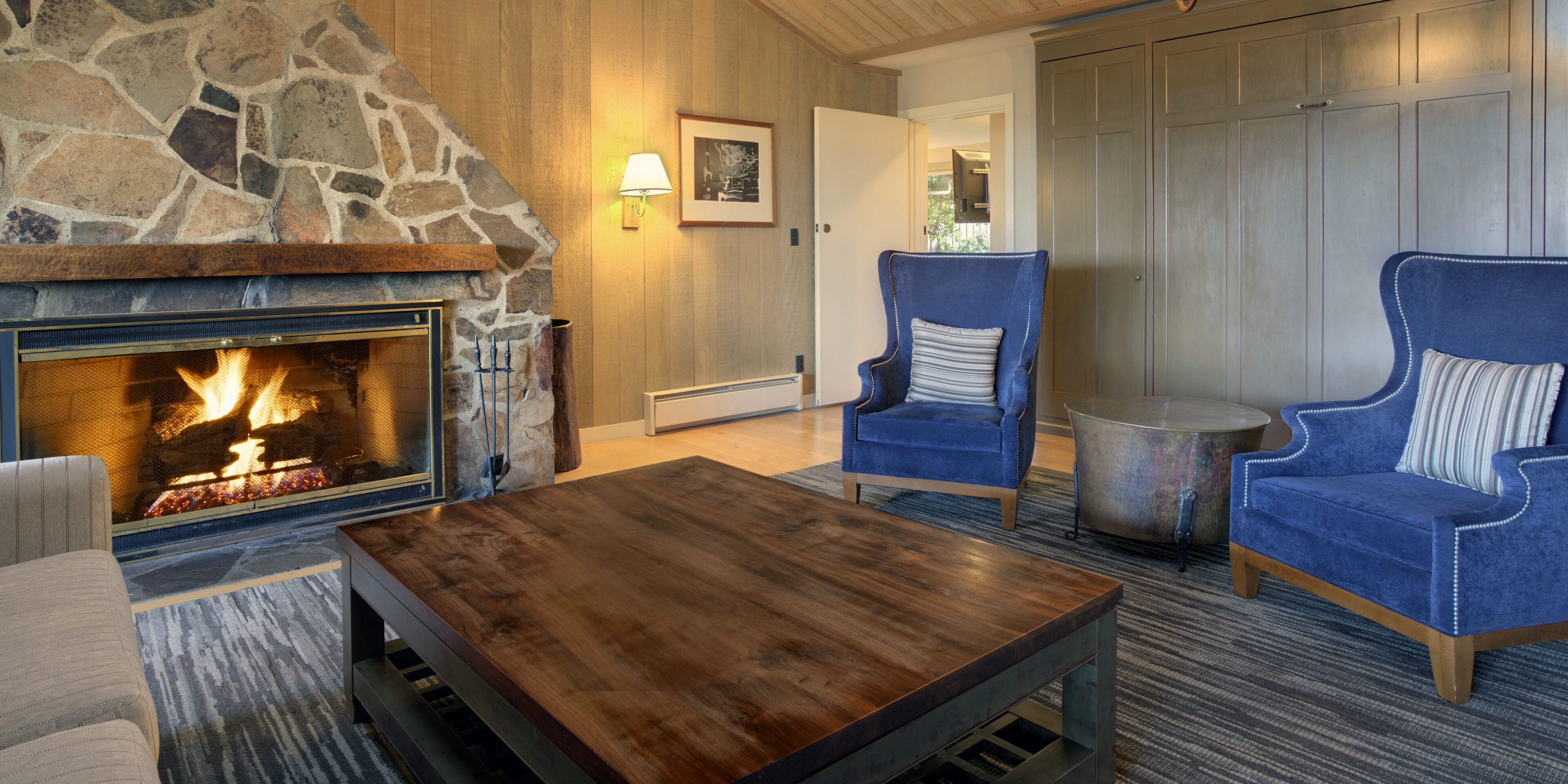 A cozy living room with a stone fireplace, two blue armchairs, a wooden coffee table, and a rug.