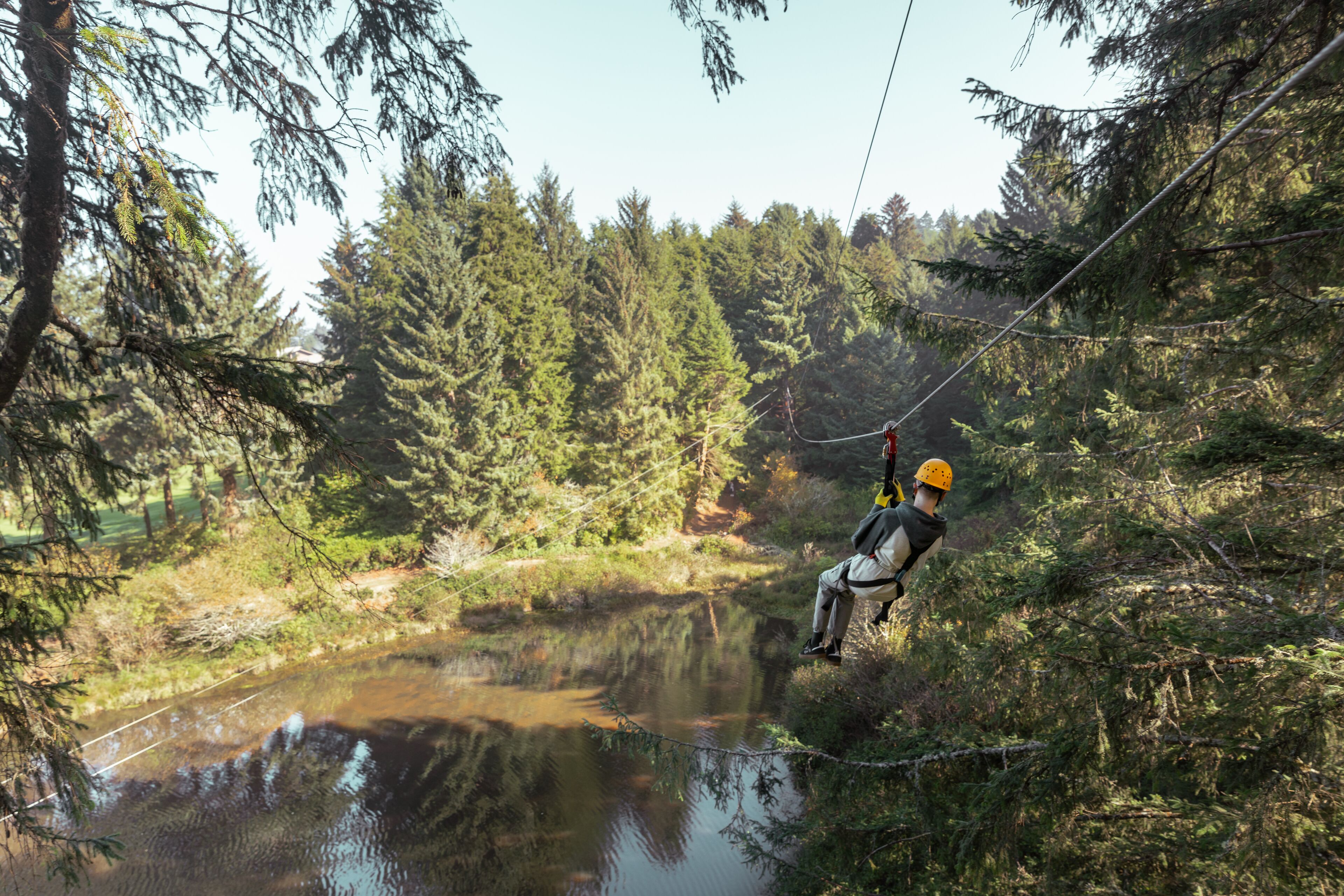En person åker på en zipline genom ett skogsområde, med en sjö eller annan vattenyta nedanför.