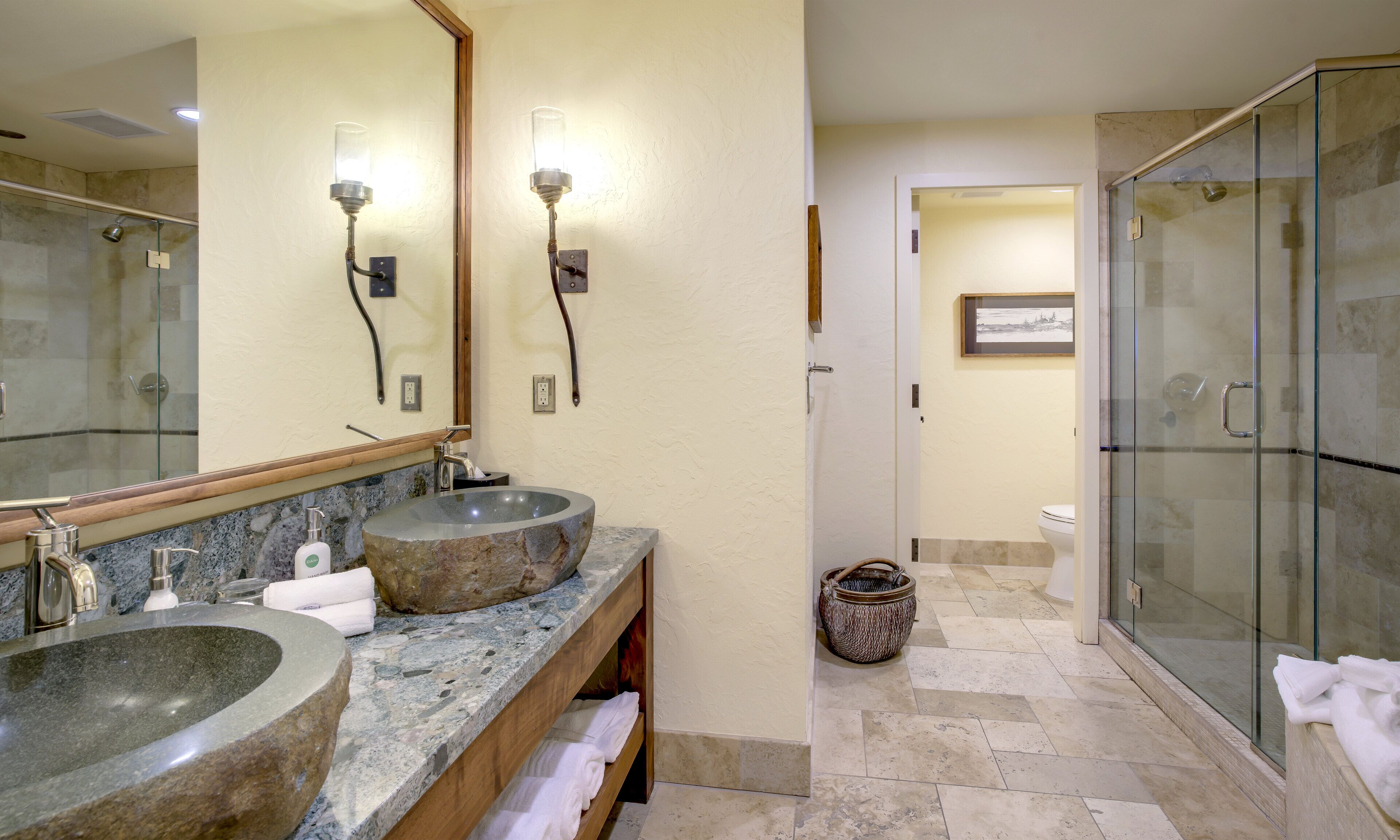A bathroom with two granite sinks, a mirror, wall sconces, a shower with glass doors, and a tiled floor.