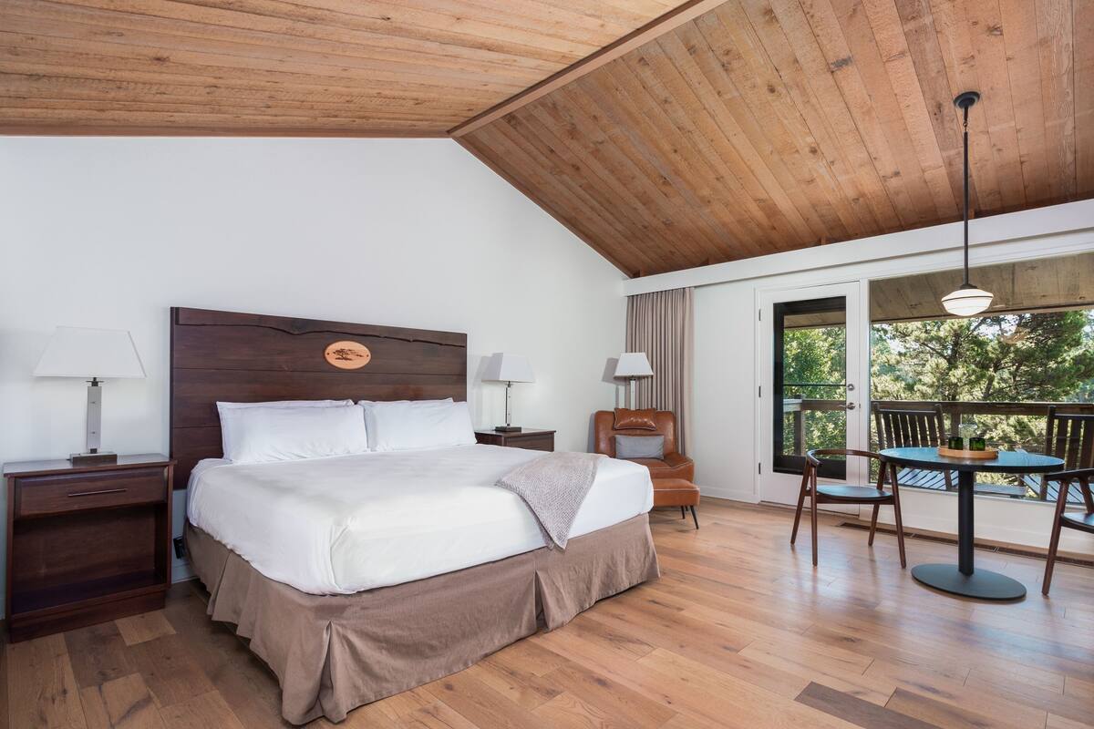 Une chambre d’hôtel avec un plafond en bois, un grand lit avec du linge de lit blanc, deux tables de chevet avec des lampes, une chaise et un balcon avec une table et des chaises.