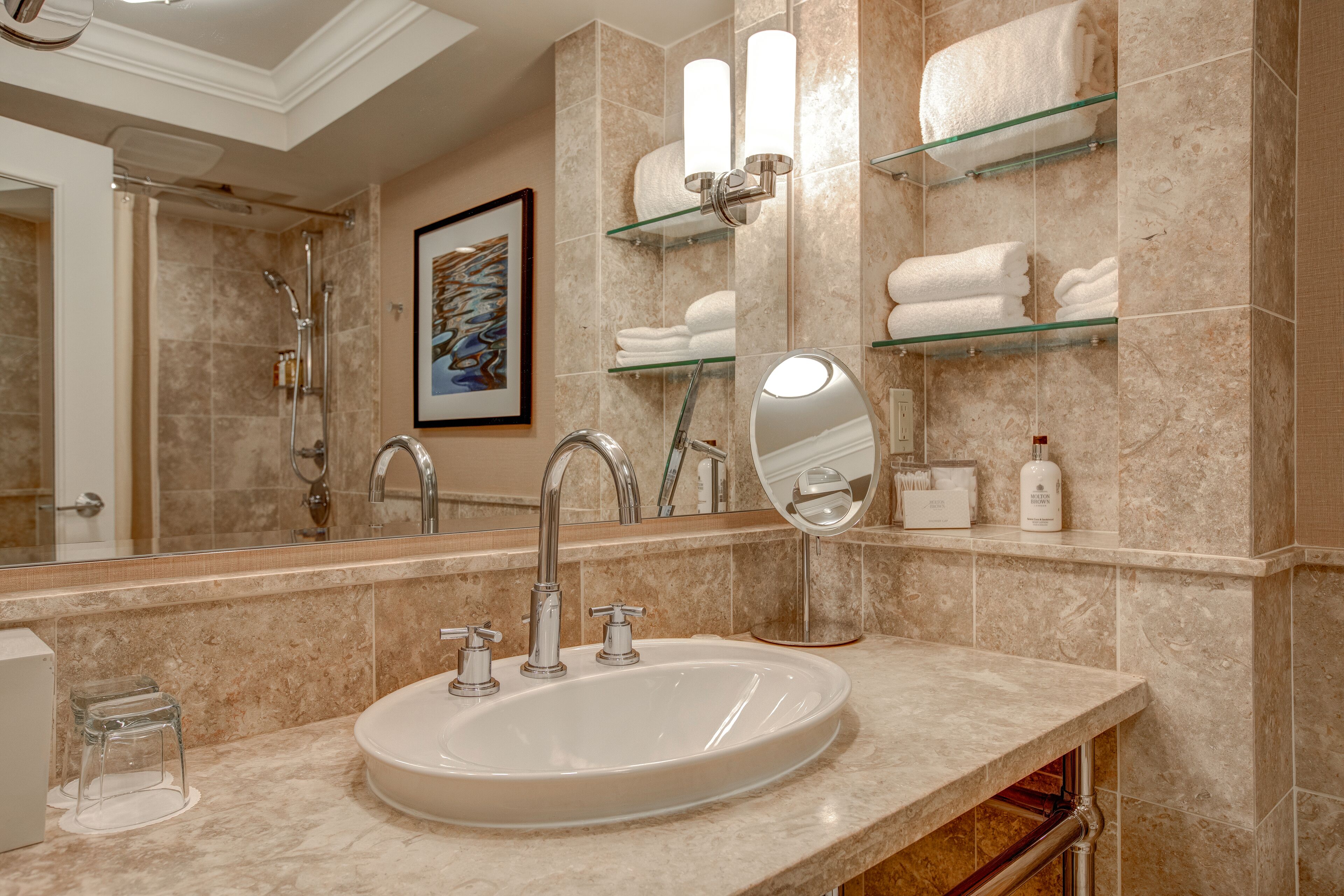 A bathroom with a sink, two faucets, a mirror, and glass shelves with towels.