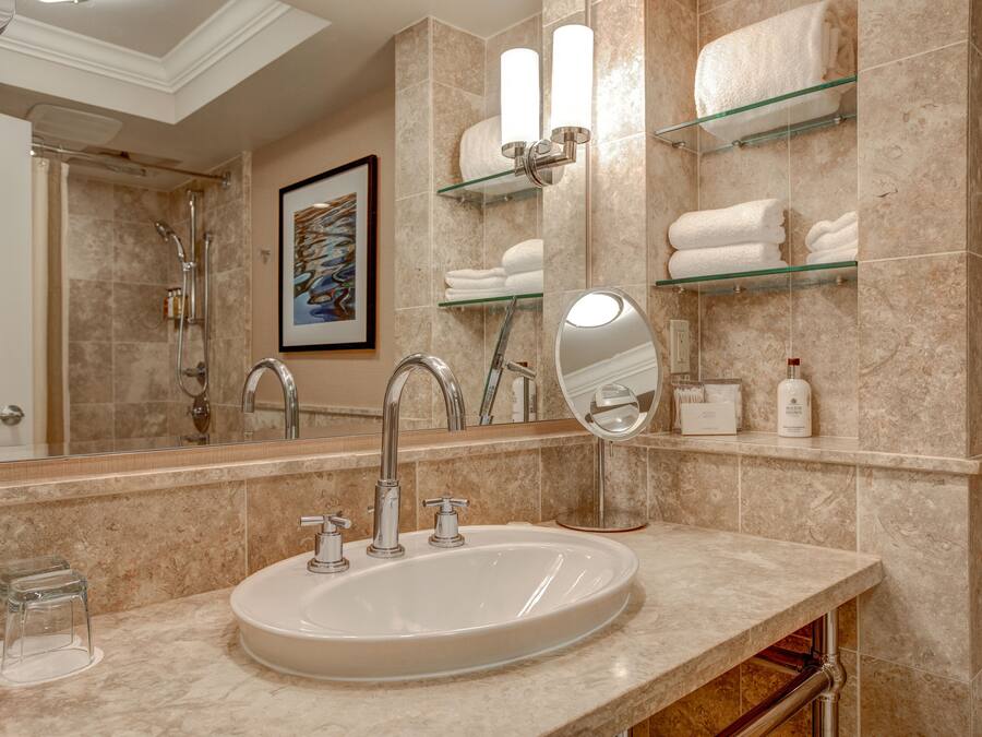 A bathroom with a sink, two faucets, a mirror, and glass shelves with towels.