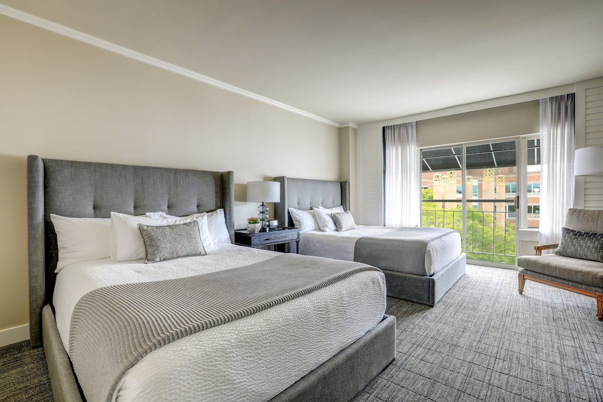 Habitación de hotel con dos camas, un escritorio con lámpara, una silla y un ventanal con cortinas.