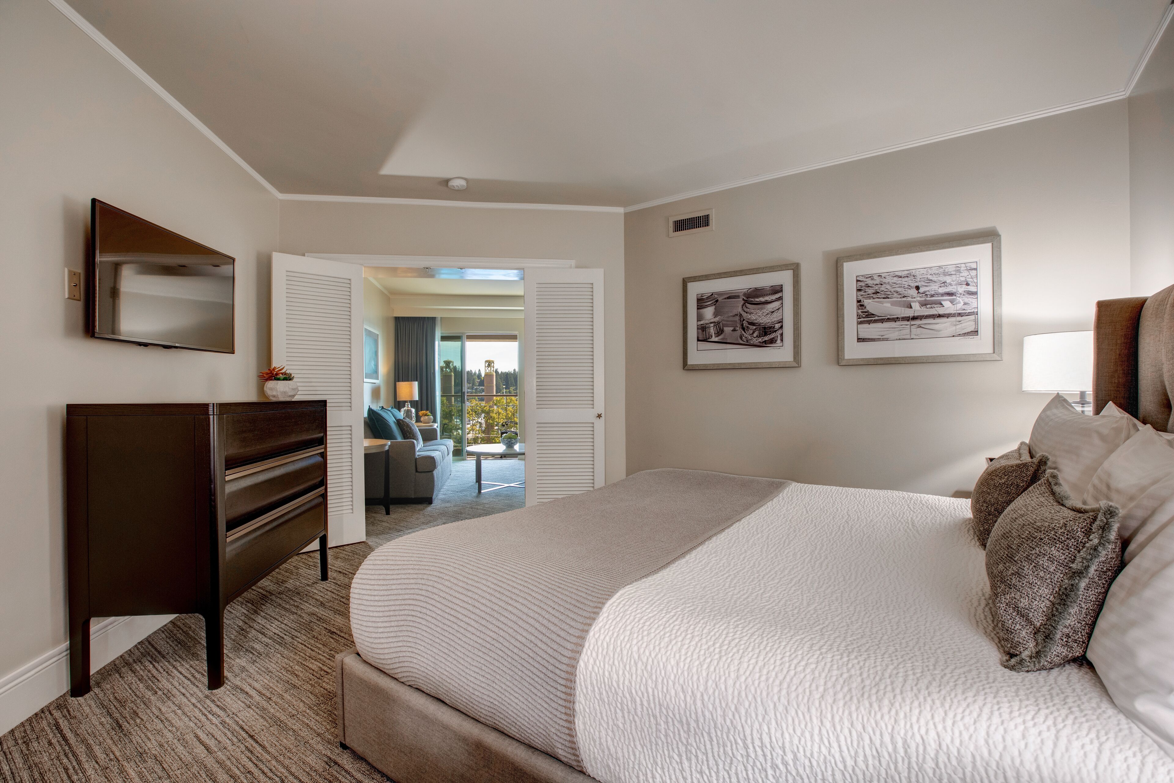 A bedroom with a large bed, a dresser, and a TV on the wall. There are framed pictures on the wall and a view of the outside through an open door.