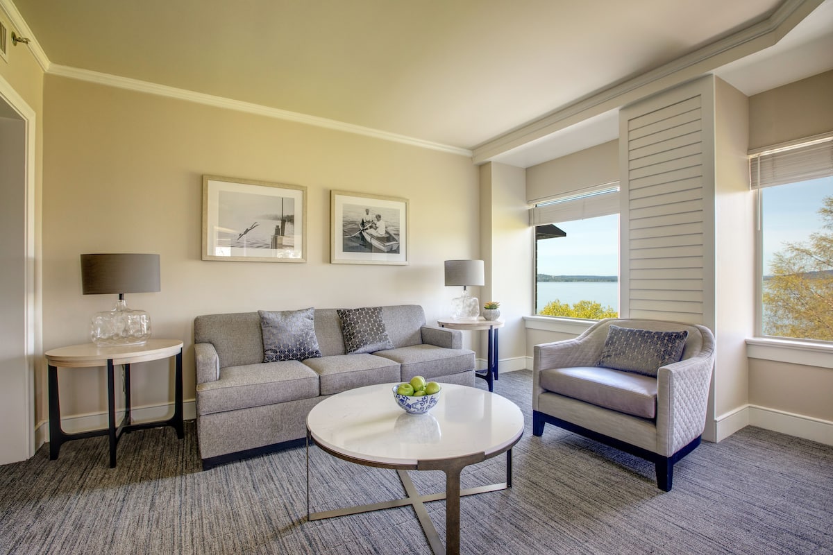 Una sala moderna con un sofá gris, una mesita redonda con un frutero, y vistas al exterior a través de grandes ventanales.