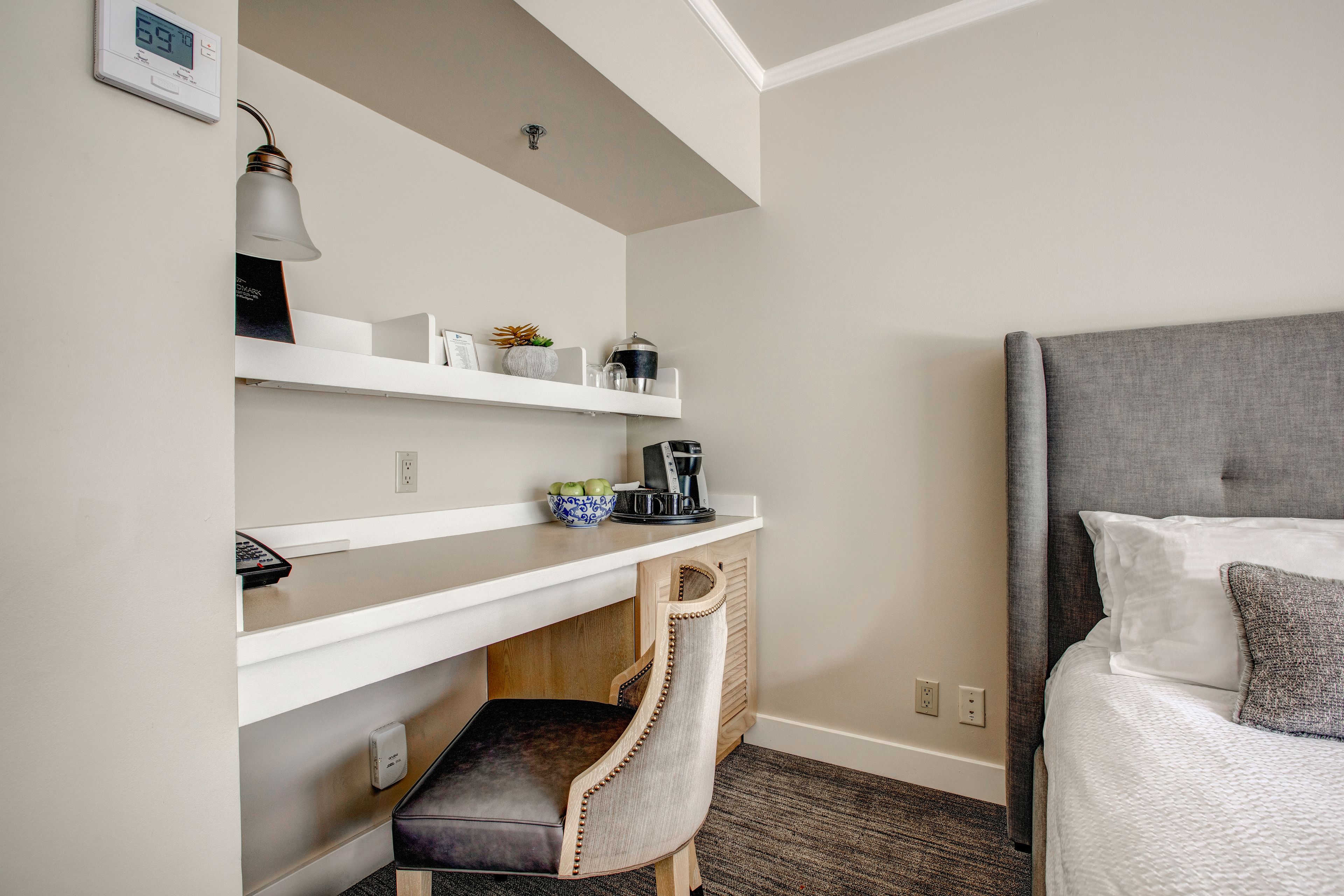 A hotel room with a bed, a desk with a lamp, a chair, and a shelf with various items.