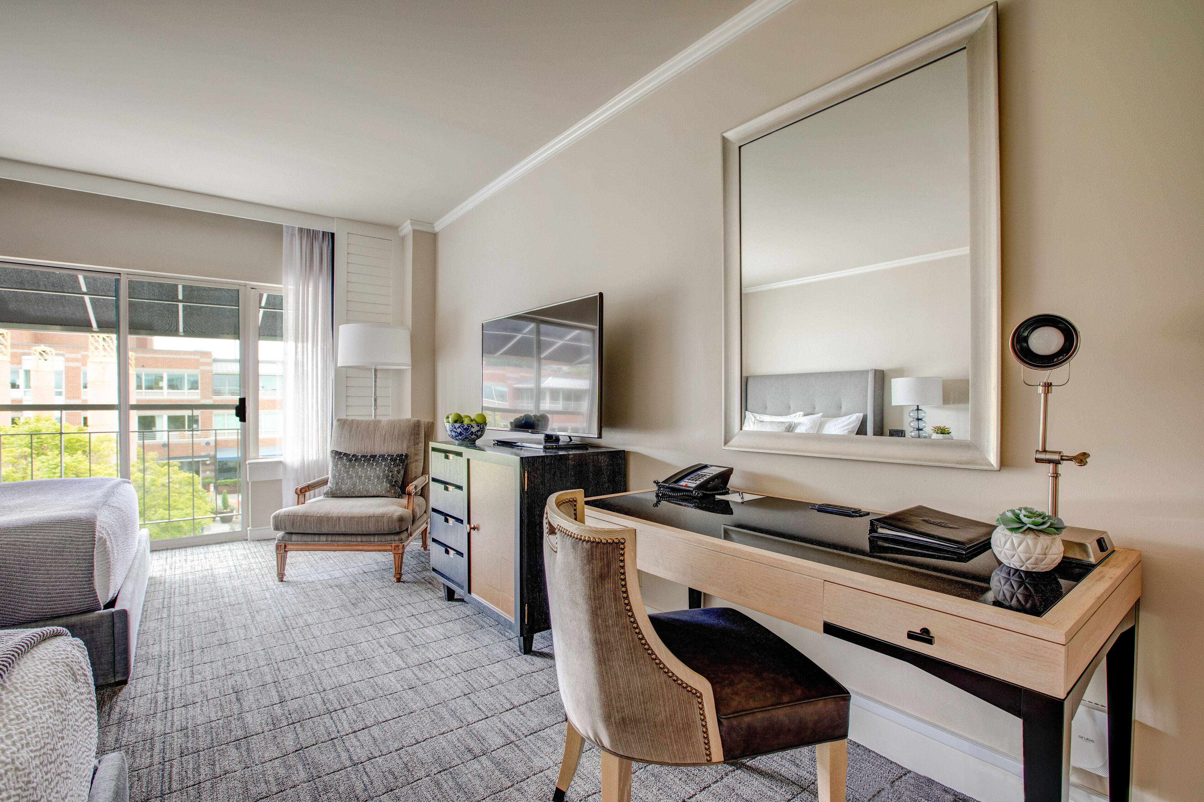 A hotel room with a large window, a desk with a computer, a chair, a bed, and a dresser.