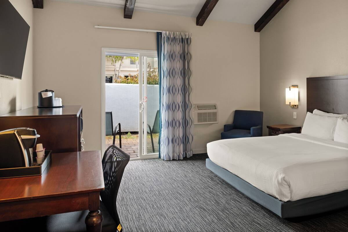 A hotel room with a bed, a desk with a coffee maker, a chair, a nightstand, and a view through a door.