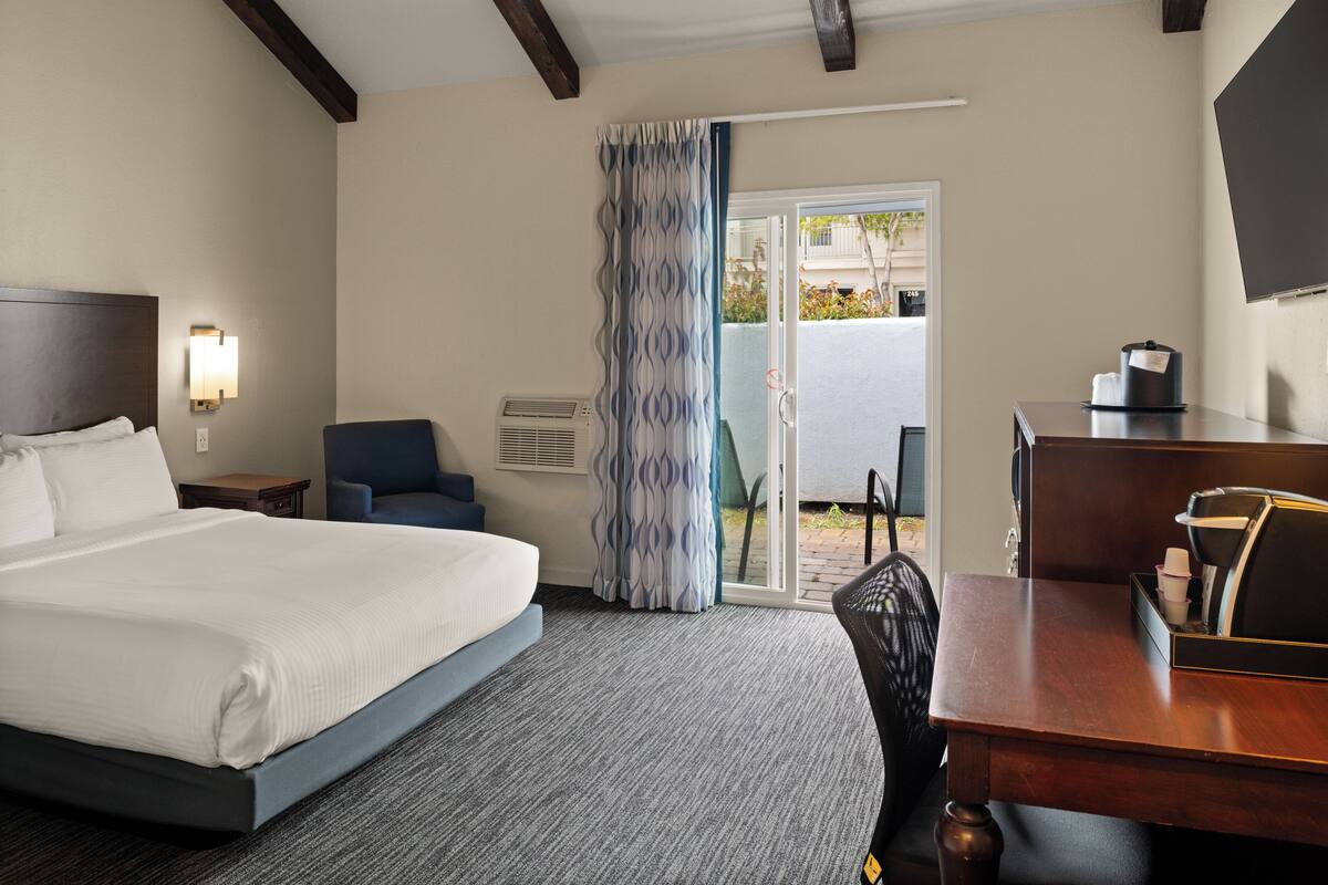 A hotel room with a bed, a desk, a chair, a curtain, and a door leading to an outdoor area.