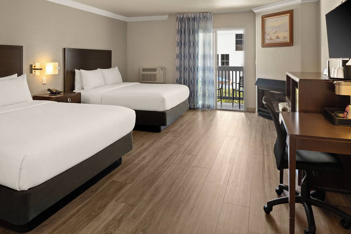 A hotel room with two beds, a desk, a chair, an air conditioning unit, and a view of the outdoors.