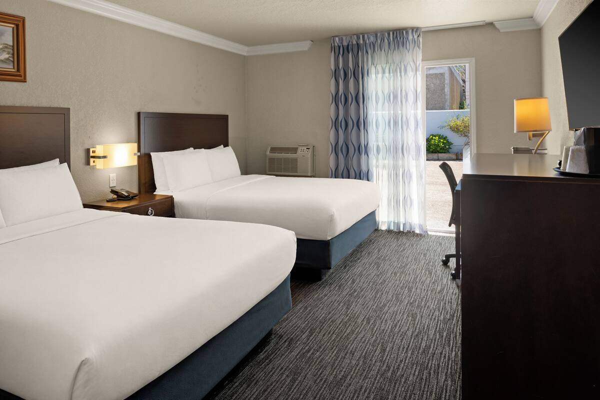 A hotel room with two beds, a desk, a chair, a lamp, and a window with curtains.