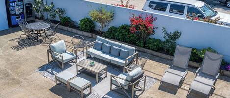 An outdoor seating area with a sofa, chairs, and a table, a vending machine, and a white wall with plants and flowers.