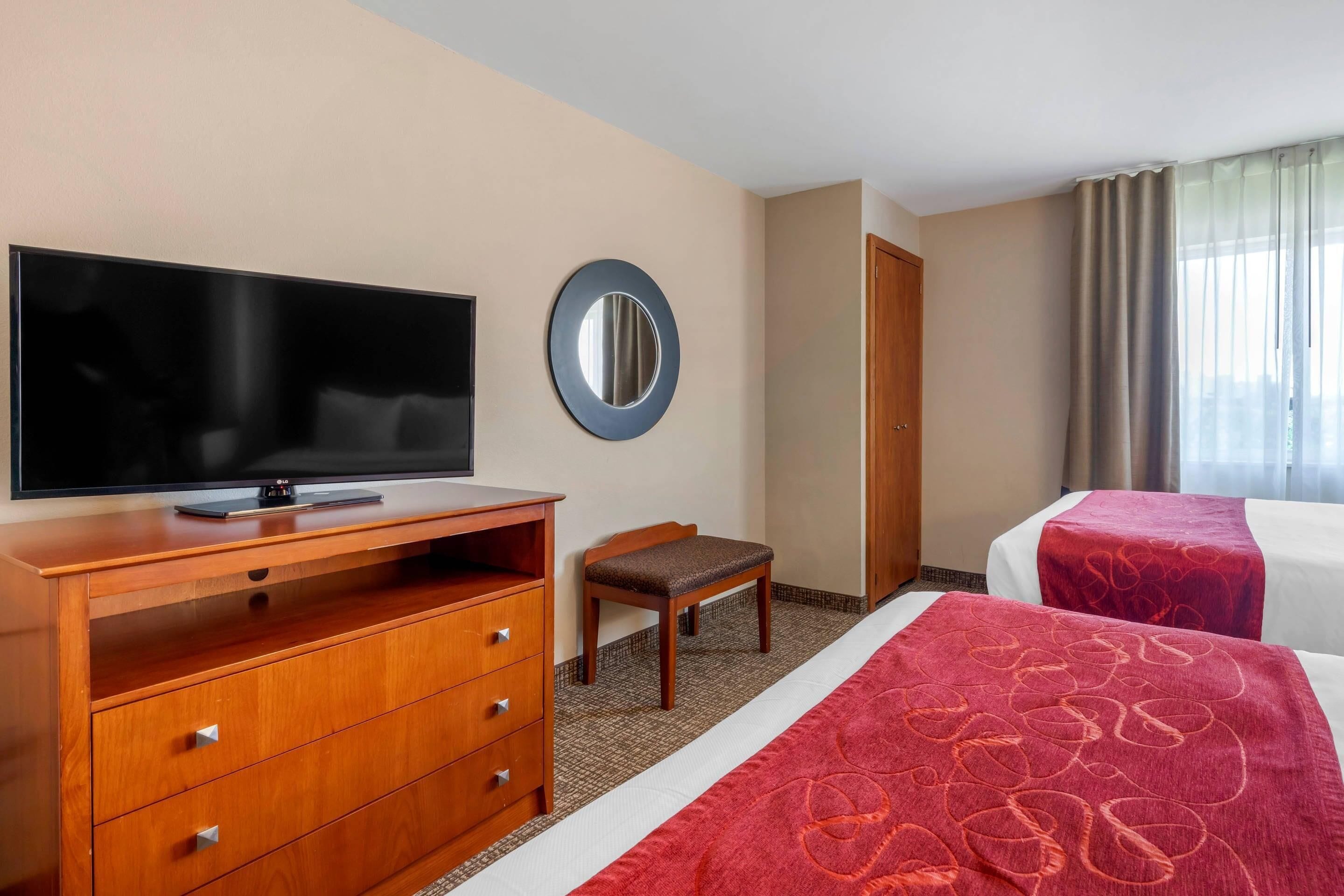 Une chambre d’hôtel avec un téléviseur posé sur une commode en bois, une petite chaise, deux lits recouverts de couvre-lits rouges, un miroir rond accroché au mur et une porte.