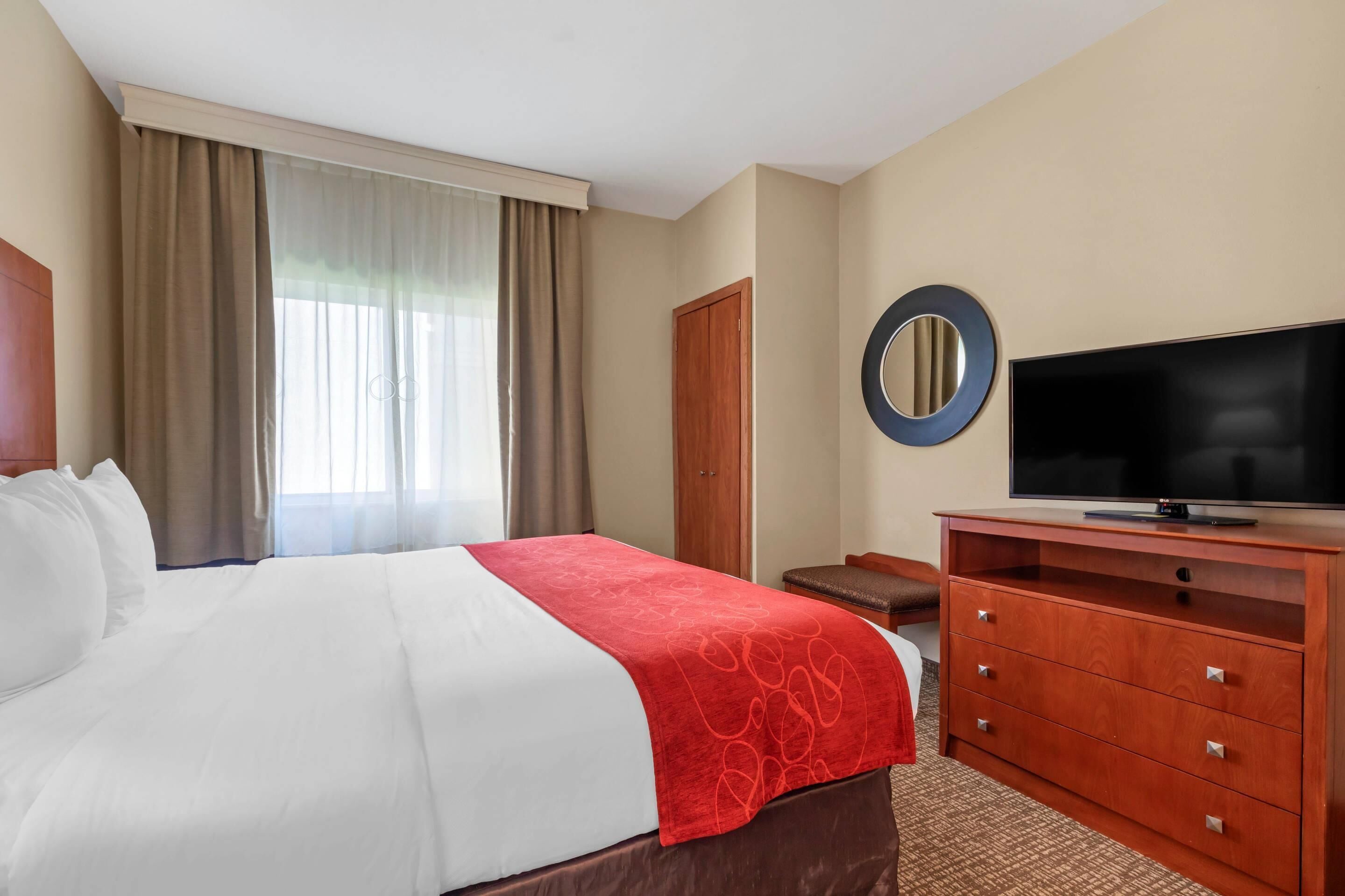 Una habitación de hotel con cama, televisor, cómoda de madera y ventana con cortinas.