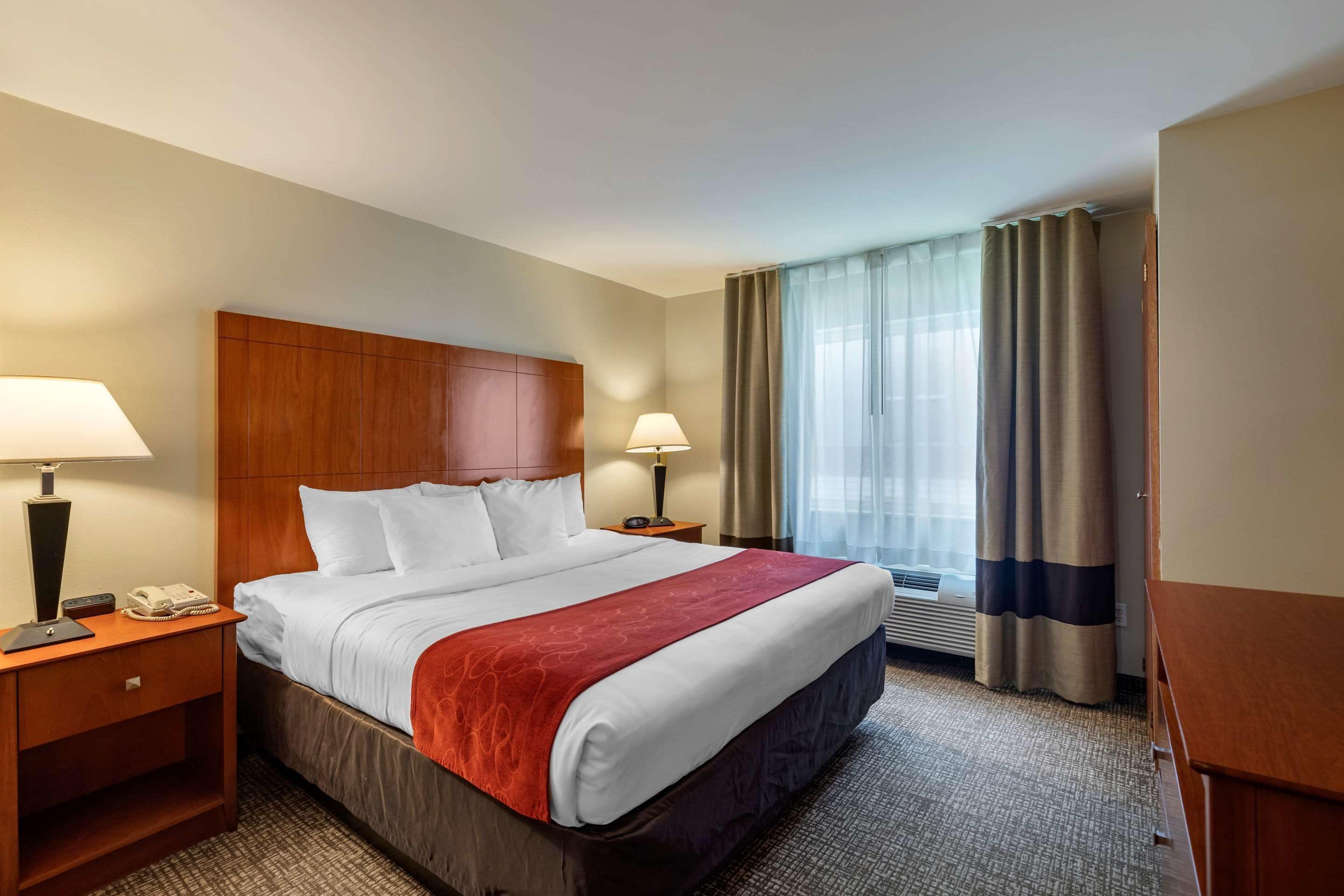 Ein Hotelzimmer mit einem großen Bett, zwei Nachttischen inklusive Lampen, einem hölzernen Kopfteil, einem Fenster mit Vorhängen und einer wandmontierten Klimaanlage.