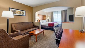 A hotel room with a sofa, a wooden table, a bed with a headboard, a desk with a lamp, a television, and a framed picture on the wall. - Comfort Suites North Dallas (Dallas)