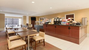 Un comedor de hotel con una larga mesa de madera y sillas, una encimera de cocina con máquinas de café y alimentos, y un amplio ventanal que proporciona luz natural.
