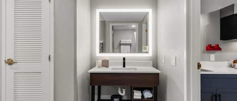 Un baño con una puerta blanca, un tocador de madera con un espejo, un lavamanos y un armario con toallas.