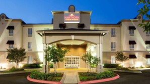 Entrada de un hotel con un vestíbulo techado, un letrero que dice "HAWTHORN SUITES BY WYNDHAM" y una zona ajardinada con árboles y arbustos.