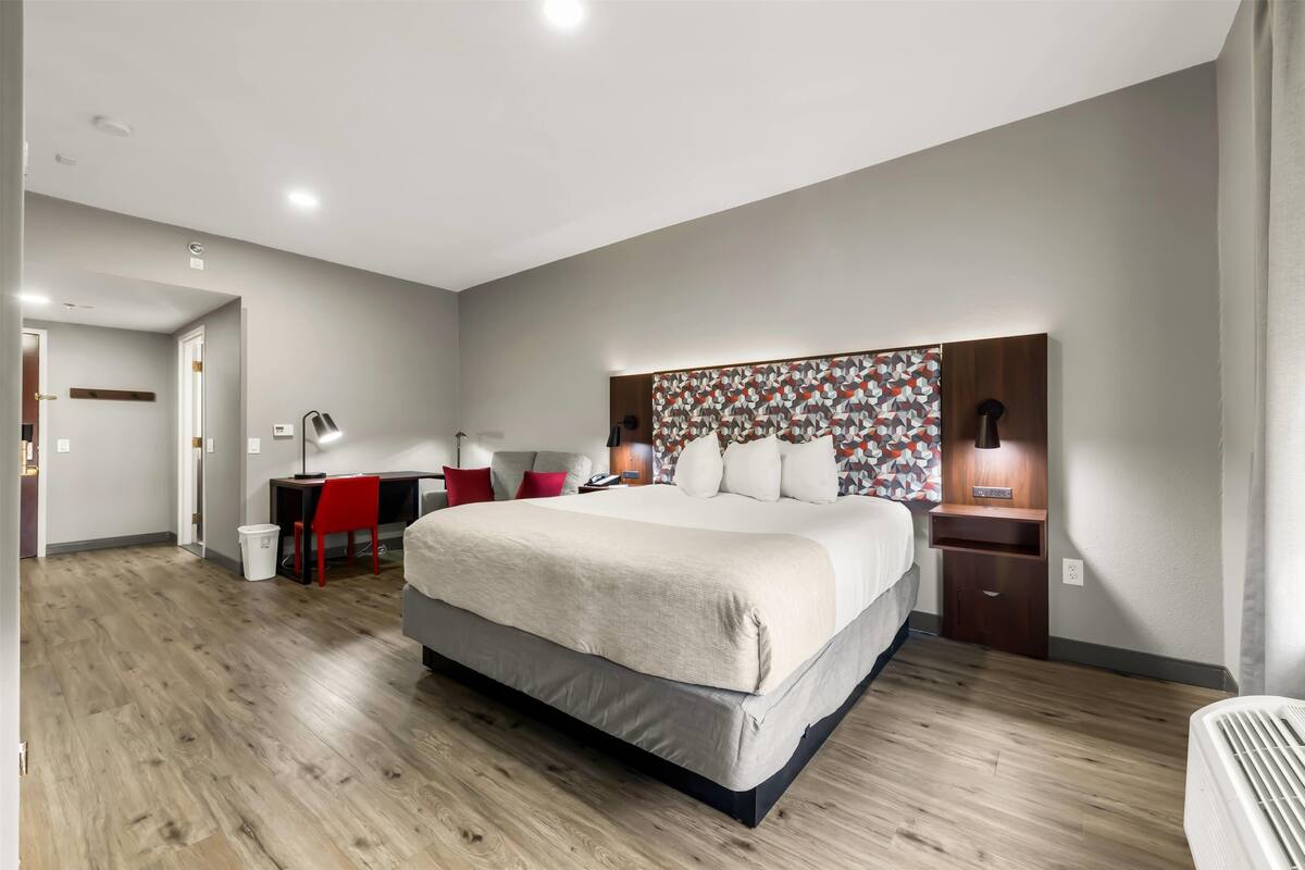 Una habitación de hotel moderna con una cama grande, un escritorio con silla, una mesita de noche con lámpara y un cabecero estampado.