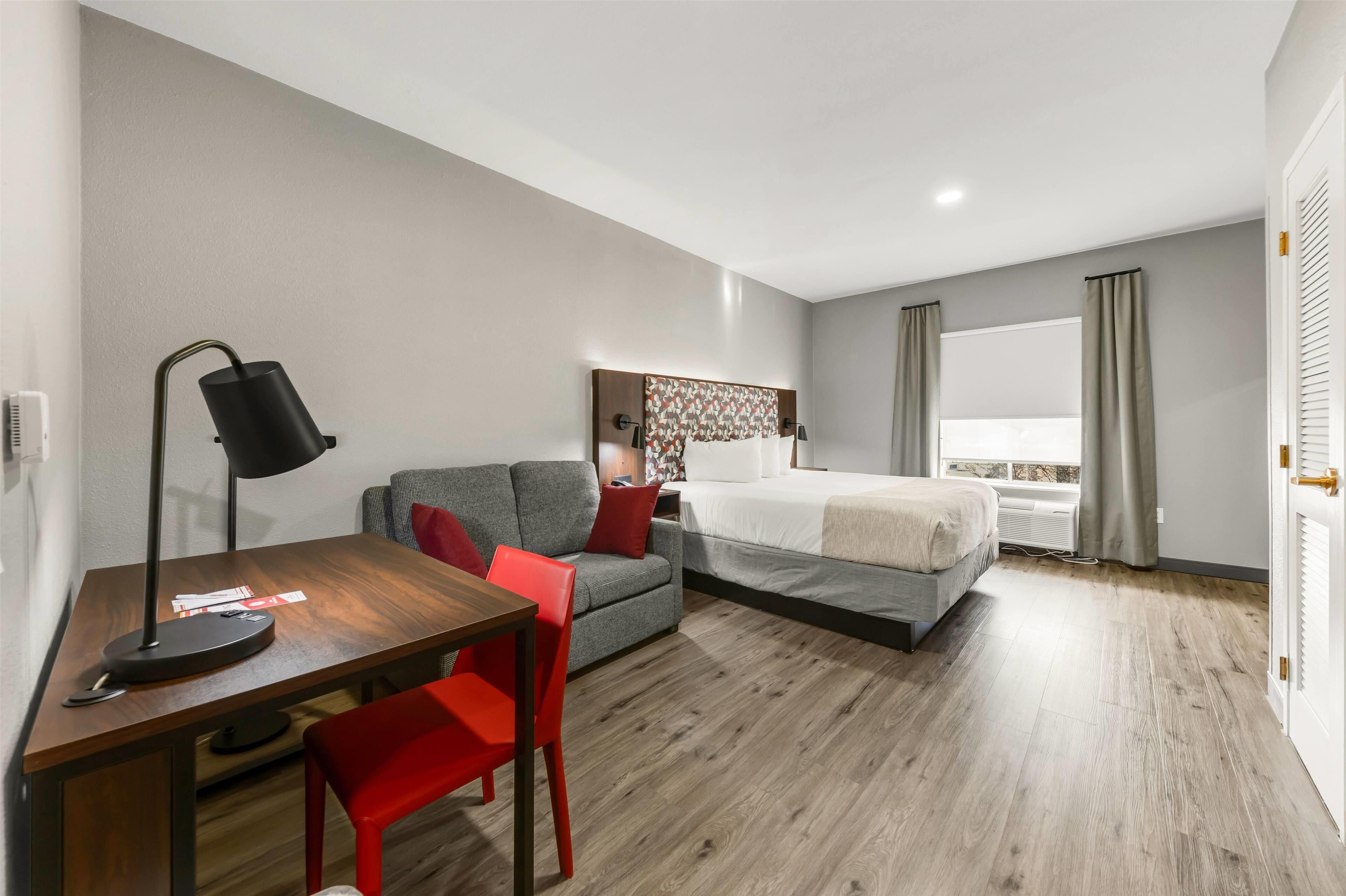 Une chambre d’hôtel avec un bureau en bois, un canapé gris, un lit et une fenêtre avec des rideaux.