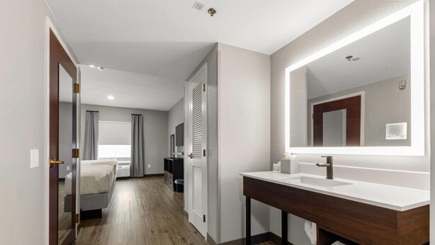 Una habitación de hotel moderna con piso de madera, un espejo grande, un lavamanos blanco y una cama con cortinas.