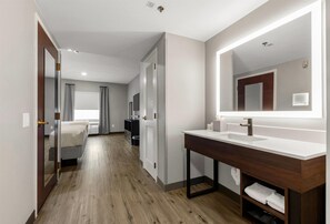 A modern hotel room with a wooden floor, a large mirror, a white sink, and a bed with curtains. - Hawthorn Extended Stay by Wyndham College Station (College Station)