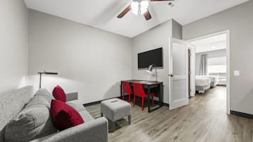 Uma sala de estar moderna com um sofá cinzento, almofadas vermelhas, um pequeno banquinho, uma escrivaninha com uma cadeira, um ventilador de teto e uma TV fixada na parede.