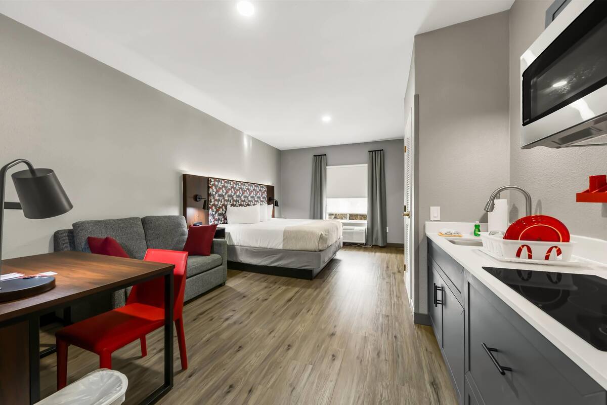 Una habitación de hotel moderna con piso de madera, un área de comedor con mesa y sillas, un sofá, una cama con cabecera, una cocina con estufa y un televisor montado en la pared.