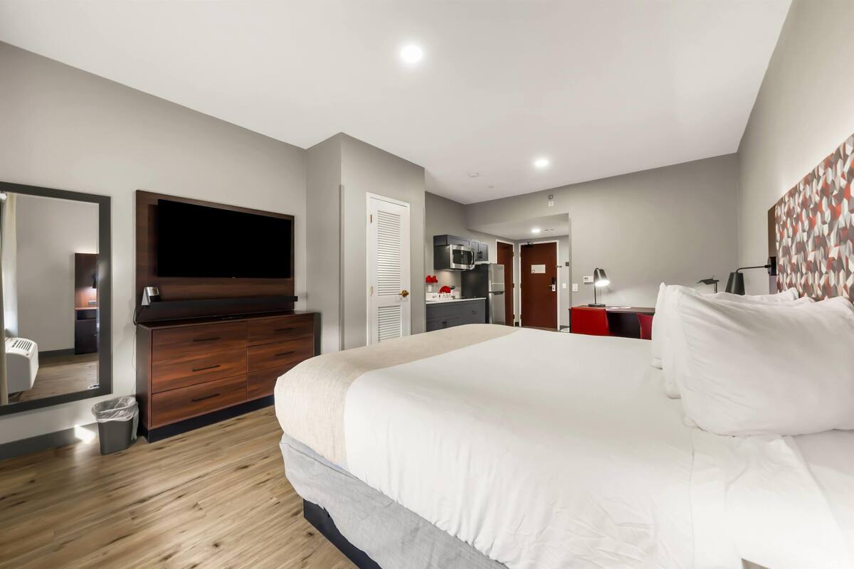 Habitación de hotel moderna con una cama grande, televisor montado en la pared, armario, una pequeña kitchenette y un baño visible a través de una puerta abierta.