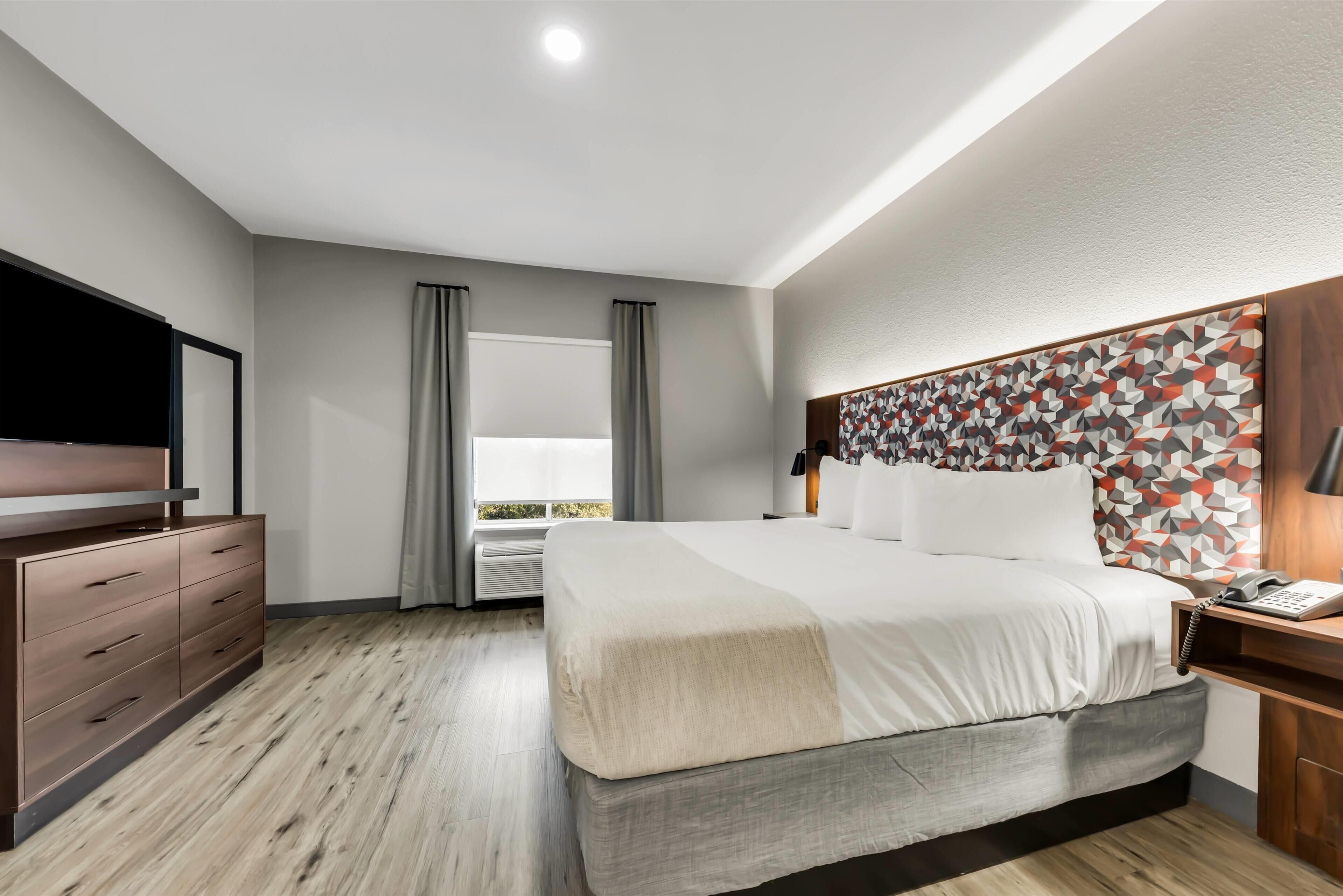 Habitación de hotel moderna con una cama grande, un aparador de madera, un televisor de pantalla plana y una ventana con cortinas.