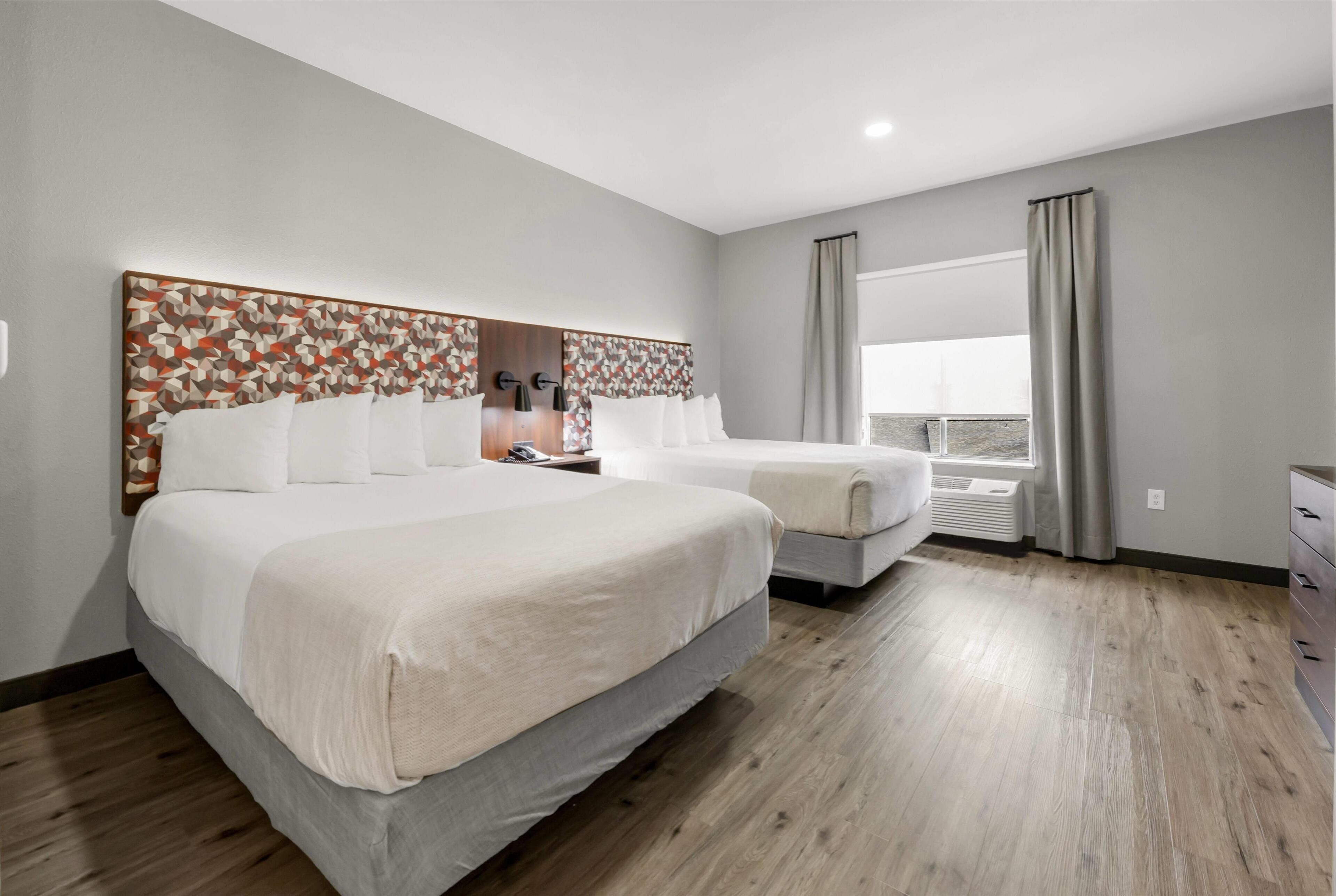 Una habitación de hotel con una cama grande, una cama más pequeña, un aparador, una ventana con cortinas y una pared con diseño geométrico.