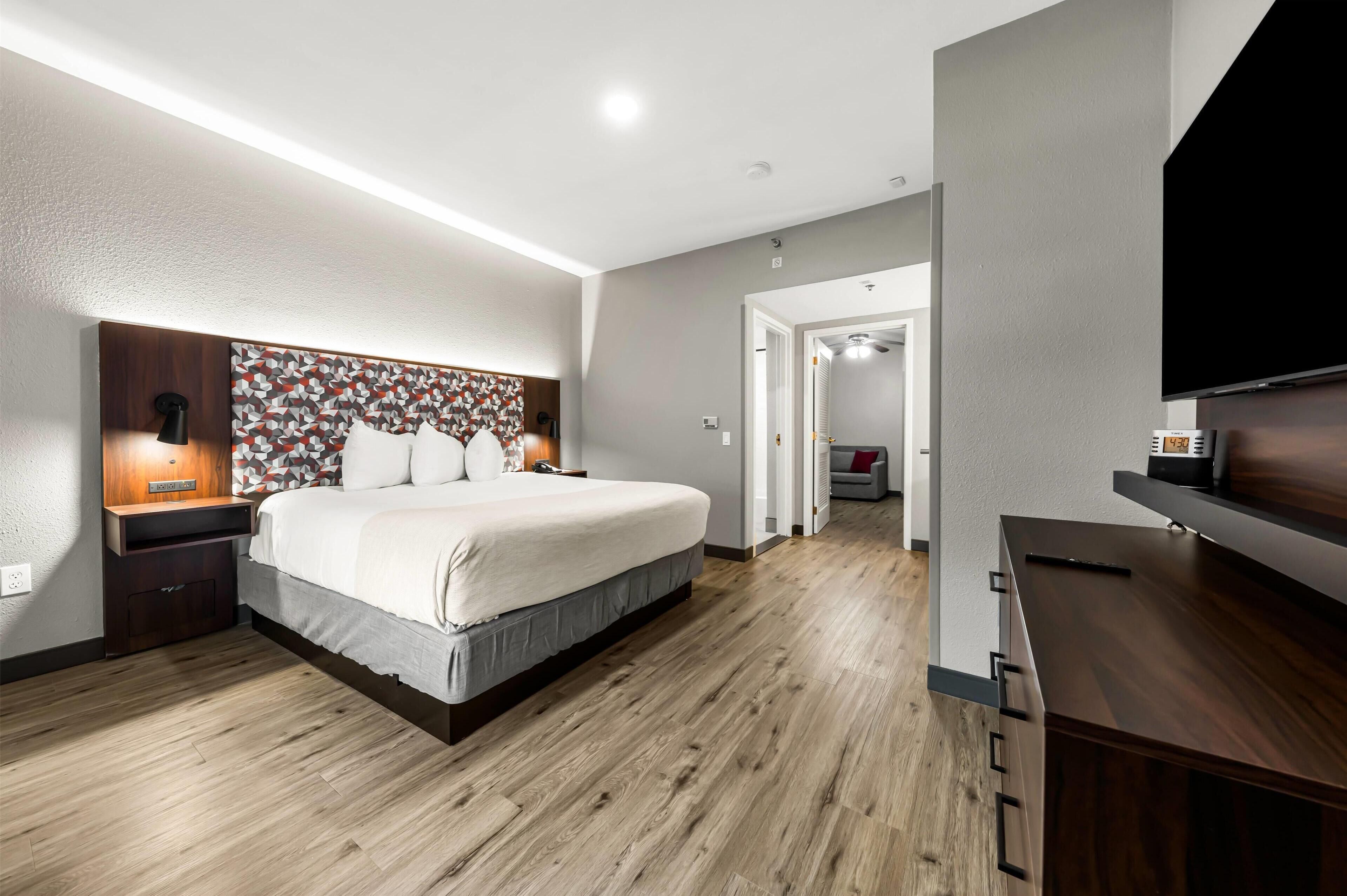 A modern hotel room with a large bed, wooden flooring, a decorative headboard, a bedside table with a lamp, and a flat-screen TV mounted on the wall.