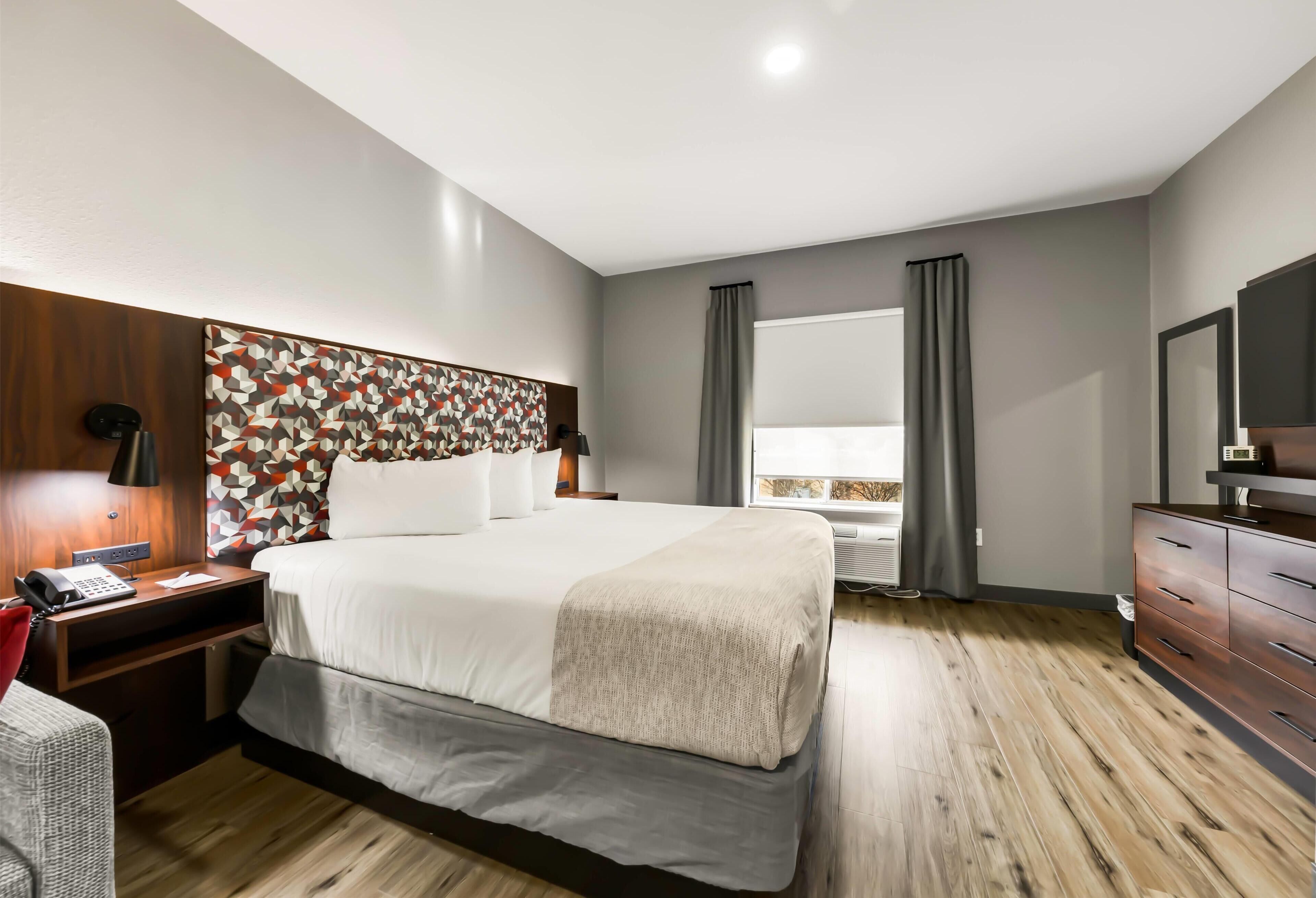 Una habitación de hotel moderna con una cama grande, cabecero de madera, mesitas de noche con lámparas, un aparador y un espejo amplio.
