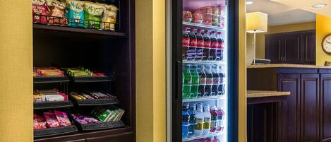 A vending machine stocked with snacks and beverages, located in a hotel lobby with wooden cabinets and a clock on the wall.