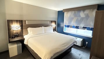 A modern hotel room with a large bed, bedside tables with lamps, a window with patterned curtains, and a built-in air conditioning unit.
