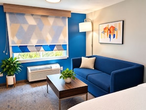 Une pièce avec un canapé bleu, une table basse en bois, une plante en pot, un radiateur et une fenêtre avec un store à motifs géométriques.