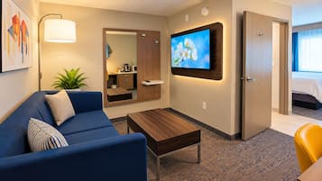 Habitación de hotel con sofá, una mesita de centro, televisor montado en la pared, un espejo y una puerta que da a un dormitorio.