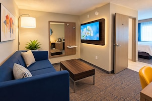 Habitación de hotel con sofá, una mesita de centro, televisor montado en la pared, un espejo y una puerta que da a un dormitorio.