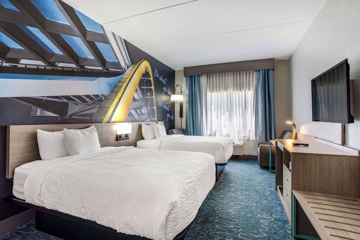 A hotel room with two beds, a desk, a TV, and a large window with curtains.